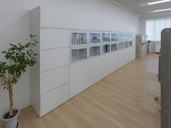 白い収納キャビネットと観葉植物を配置した明るいオフィスの通路。木目フロアが続くシンプルな職場インテリア。