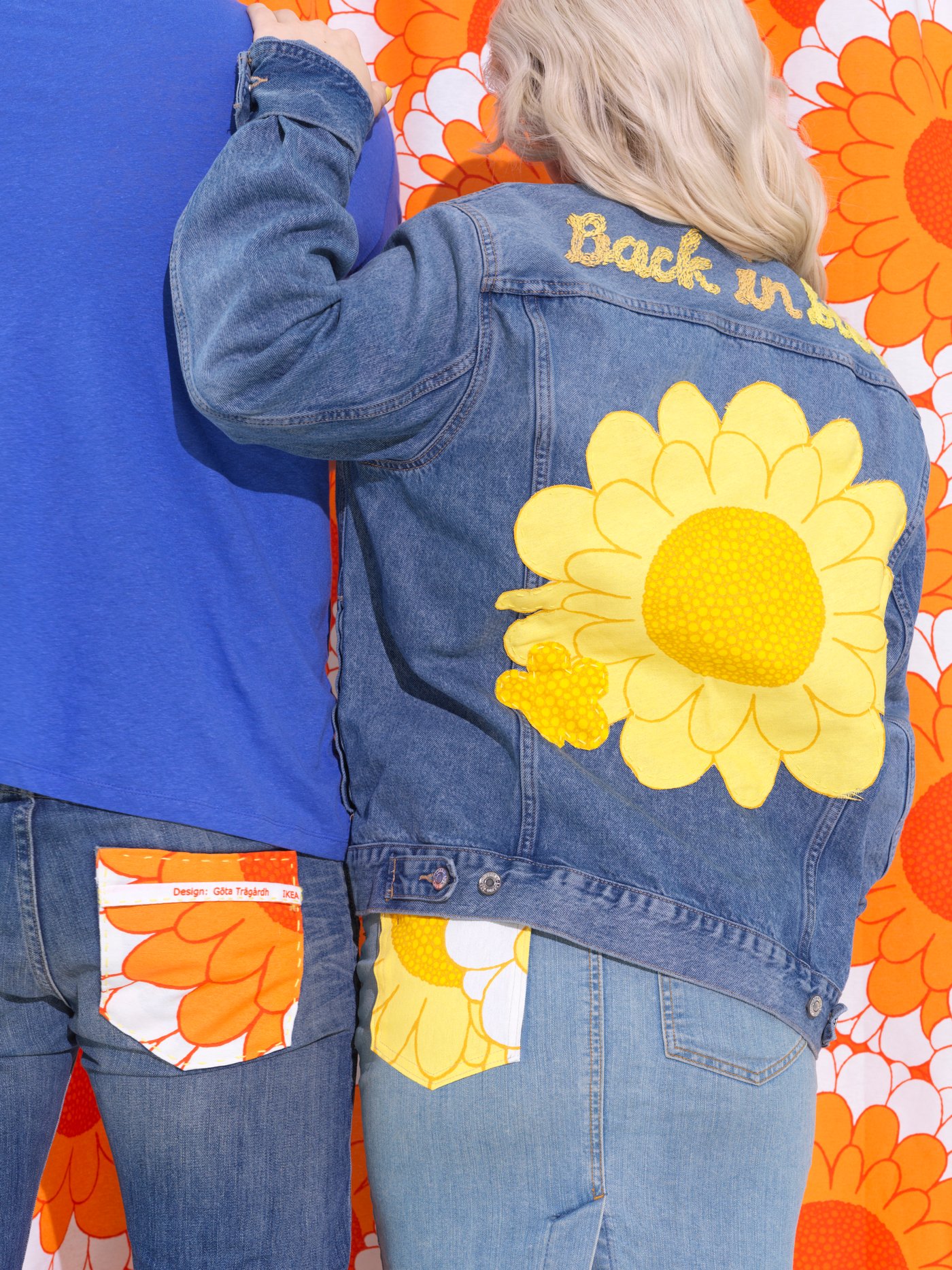 Dua orang kelihatan di belakang. Kedua-dua mereka memakai pakaian denim, dengan sedikit fabrik berbunga-bunga SANDETERNELL berwarna jingga atau kuning.