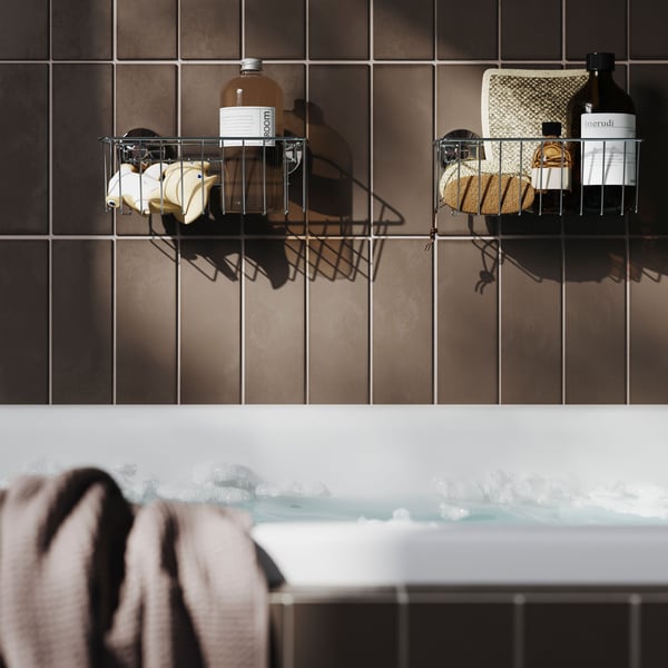 Two zinc-plated KROKFJORDEN baskets with suction cups are filled with toiletries on a brown-tiled wall above a bathtub.