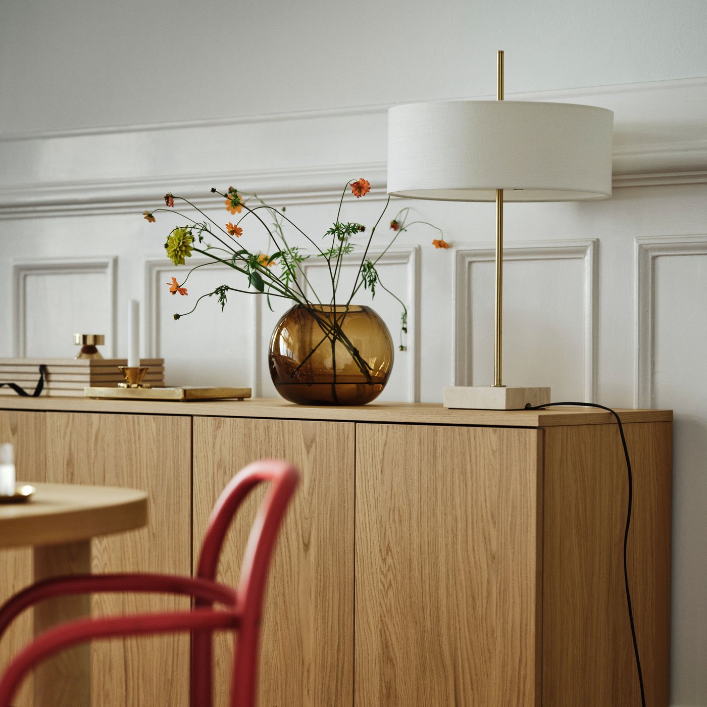 A brass-plated and white STOCKHOLM 2025 table lamp sits next to a brown vase on a cabinet by a white wall with wooden panels.