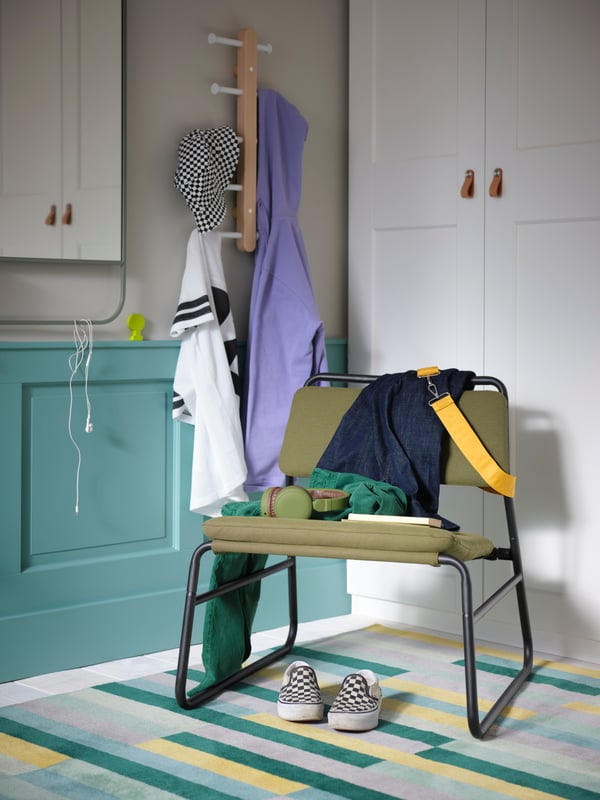 Un fauteuil LINNEBÄCK vert olive sur un tapis multicolore, à côté d’un miroir mural vert et d’une patère à laquelle sont pendus des vêtements.