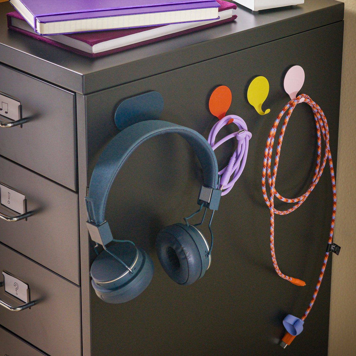 A black HELMER drawer unit with multicolour GRESLA hooks with magnets attached, holding earphones and chords.