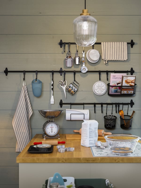 A kitchen island with bakeware, black rails and hooks with baking tools hanging on them against a green wooden wall.