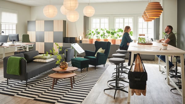 Bild på ett kontor som är indelat i olika zoner. Till vänster syns kontorsbord och kontorsstolar, i mitten finns ett loungeutrymme med soffa och fåtölj och till höger finns ett konferransbord där två personer sitter på vars en pall och pratar.