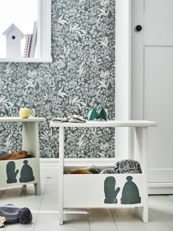 Two PERJOHAN stools with storage, painted in pale colours and with a hat and a mitten painted on them, stand in a hallway.