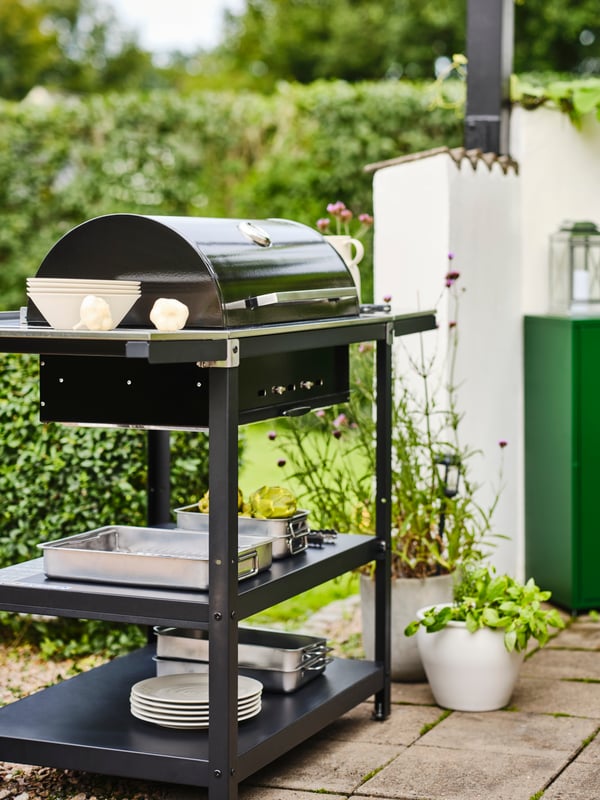 Terasa s GRILLSKÄR roštiljem na ćumur s 2 pomoćna stočića na popločanom podu i zelenim žbunjem u pozadini.