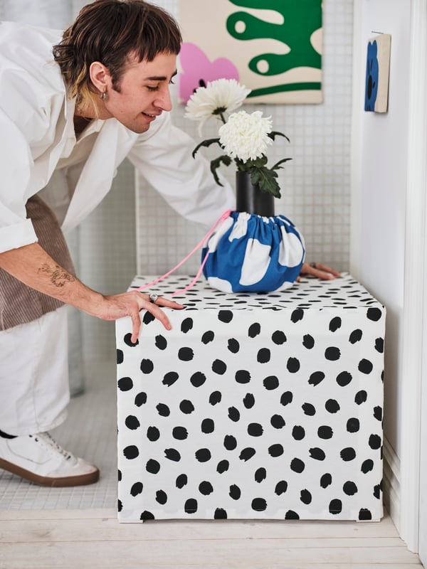 A person adjusts a small side table covered with the SANDLUSERN pre-cut fabric. On top of the table is a fabric-covered vase.