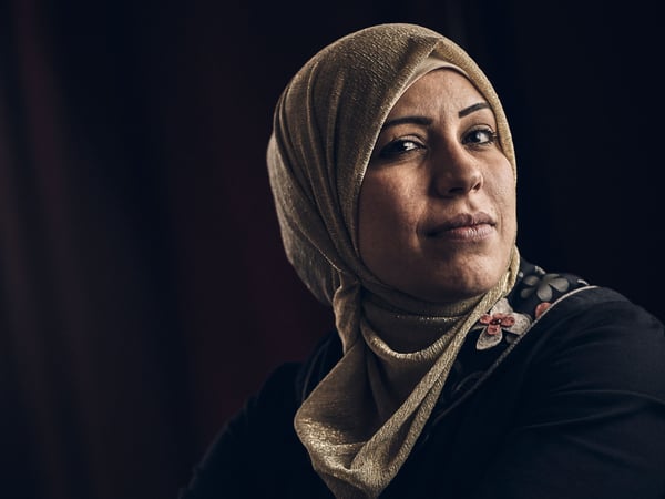 A middle eastern woman wearing a black top with an embroidered collar, and a beige hijab, against a black background.