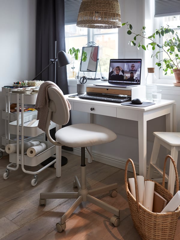 A white IDANÄS table in front of two windows set up for work with a laptop on a monitor stand on it and work chair in front.