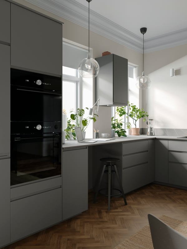 A big kitchen with dark grey VOXTORP cabinets, a wall cabinet between two windows, a brown wooden floor and black appliances.