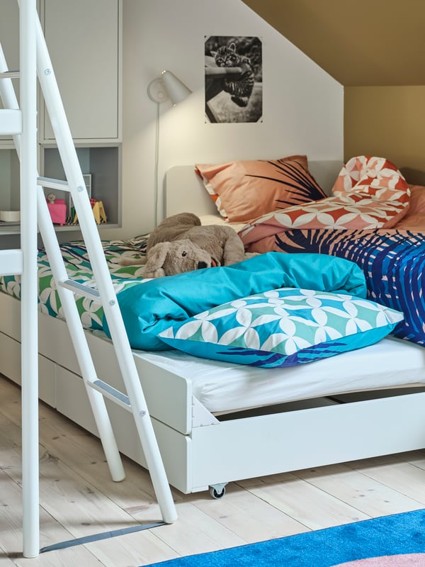 A pulled out SLÄKT underbed with GRACIÖS bed linen and a soft toy dog sits next to the ladder of a VITVAL loft bed.