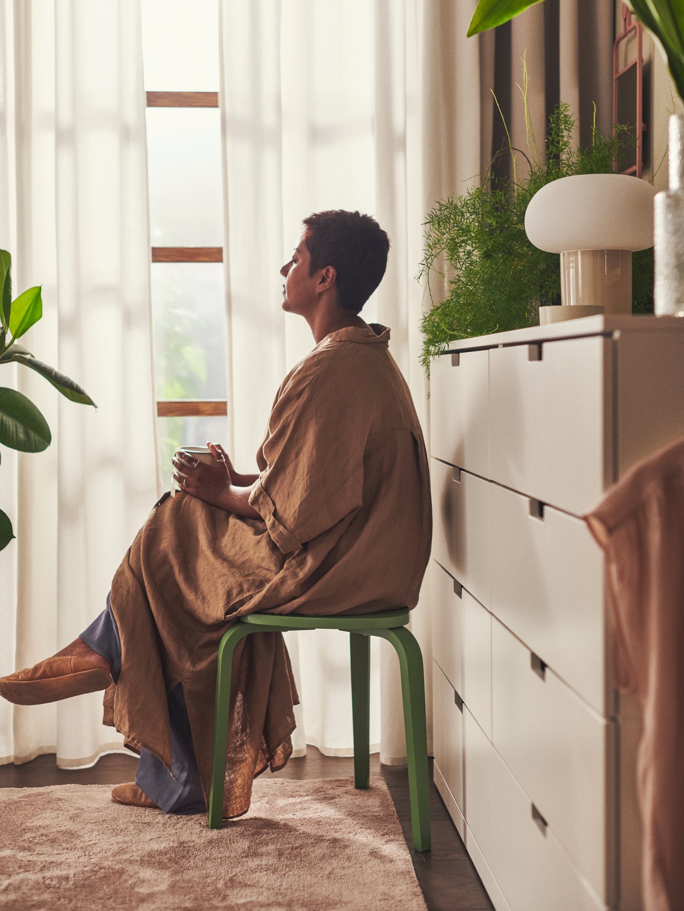 Person sitter på en grön pall vid ett fönster med ljusa gardiner, omgiven av växter och en vit NORDLI byrå med lampa och inredningsdetaljer.