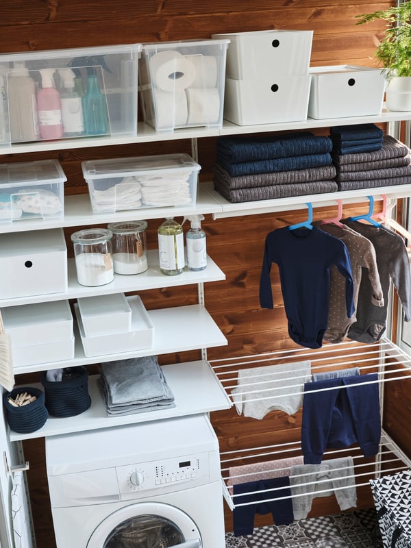 A white storage system with lots of shelves, two drying racks and a clothes rail where baby clothes hang to dry.