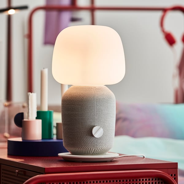 Una lámpara parlante Wi-Fi SYMFONISK de color blanco al centro de la imagen, sobre un mueble metálico rojo y frente a un plato con velas apagadas. 