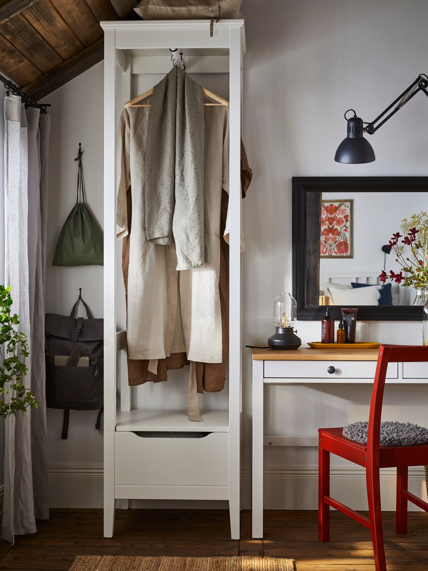 A white IDANÄS open wardrobe with clothes hanging from BUMERANG coat-hangers stands beside a HEMNES desk with 2 drawers.