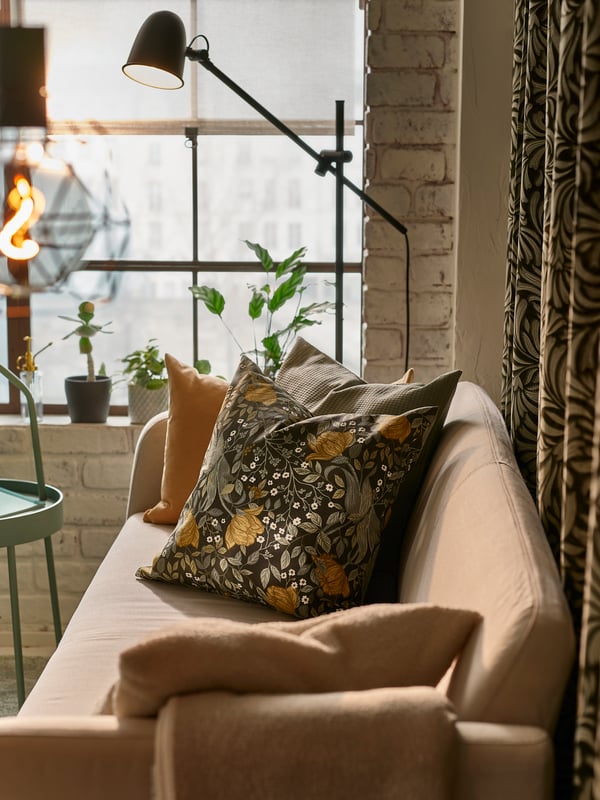 Un lampadaire noir à côté d’un canapé beige garni d’un coussin dans une housse TUVSÄV vert profond/motif floral et d’autres coussins.