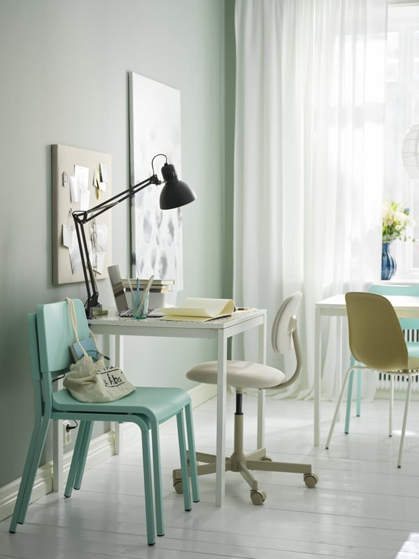 A white MELLTORP table placed by a wall, a TERTIAL work lamp on it and stacked, light turquoise TEODORES chairs next to it.