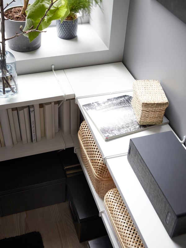 A corner where wall-mounted white shelves of different depths meet. Books, storage boxes and a speaker on the shelf.