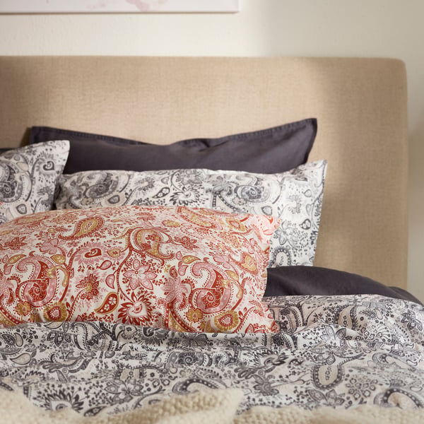 Cabeceira de cama com dois tipos de roupa de cama RODGERSIA com padrão de cornucópias, em cinzento e em vermelho.