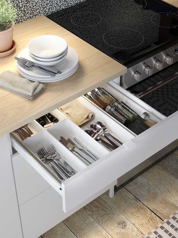 Un módulo de cajones de cocina ENHET blanco con el cajón superior abierto para mostrar una bandeja para cubiertos UPPDATERA con cubiertos y utensilios.