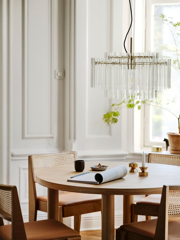 Runt matbord med fyra trästolar under en elegant STOCKHOLM 2025 taklampa i glas, med ljusinsläpp från ett fönster och grön växt i bakgrunden.