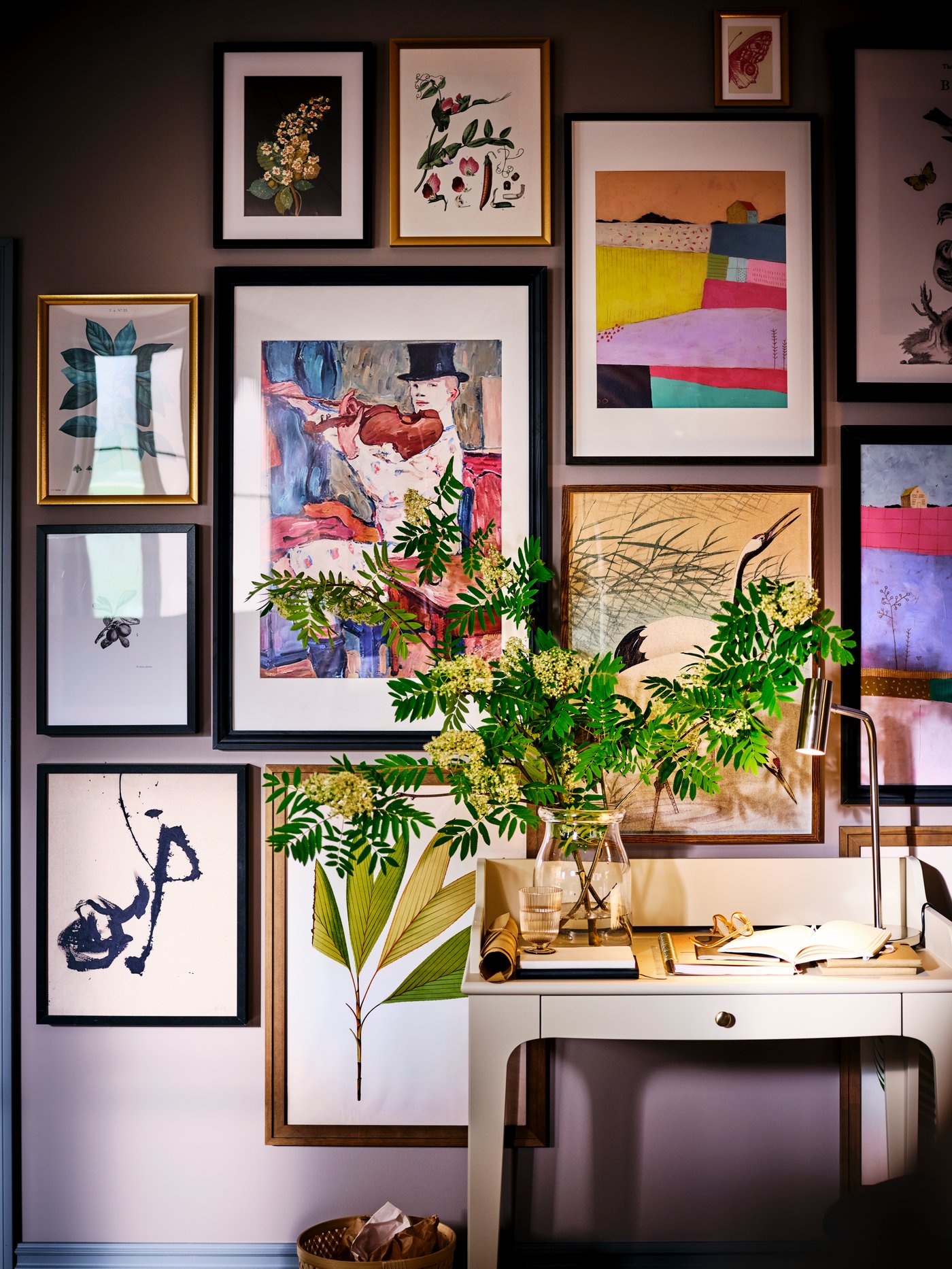 A clear glass BEGÄRLIG vase filled with flowers on a LOMMARP desk, with brightly coloured framed prints on the wall behind.
