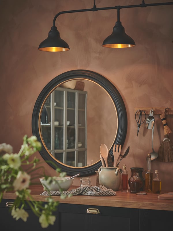 A black ALMARÖD mirror hung on an apricot-coloured wall in a rustic style kitchen, above the kitchen worktop.