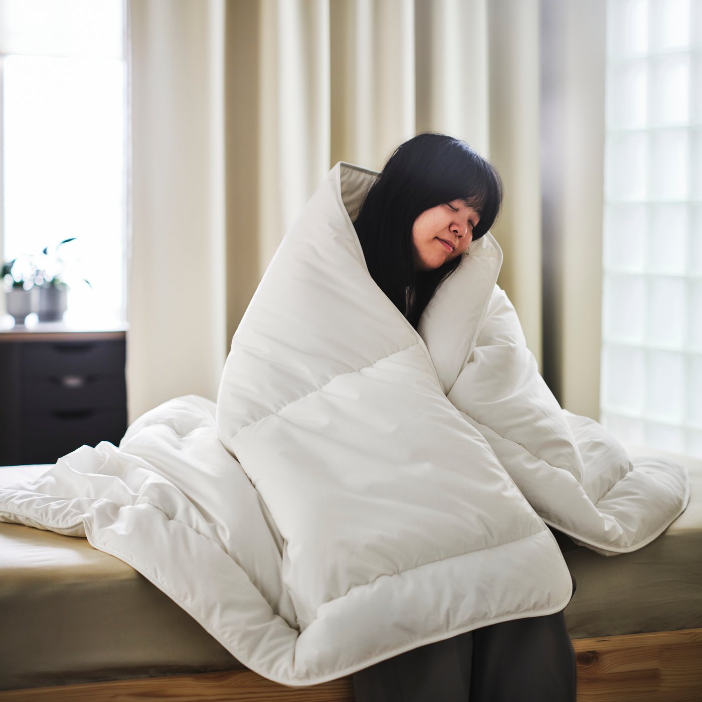 Une personne assise sur un lit couvert d’un drap beige clair, enveloppée dans une couette SKOGSFRÄKEN sans housse.