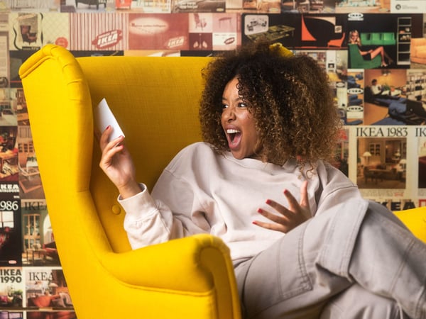 A woman with curly hair, wearing a light-colored sweatshirt and pants, sits in a yellow armchair and looks surprised at her phone.
