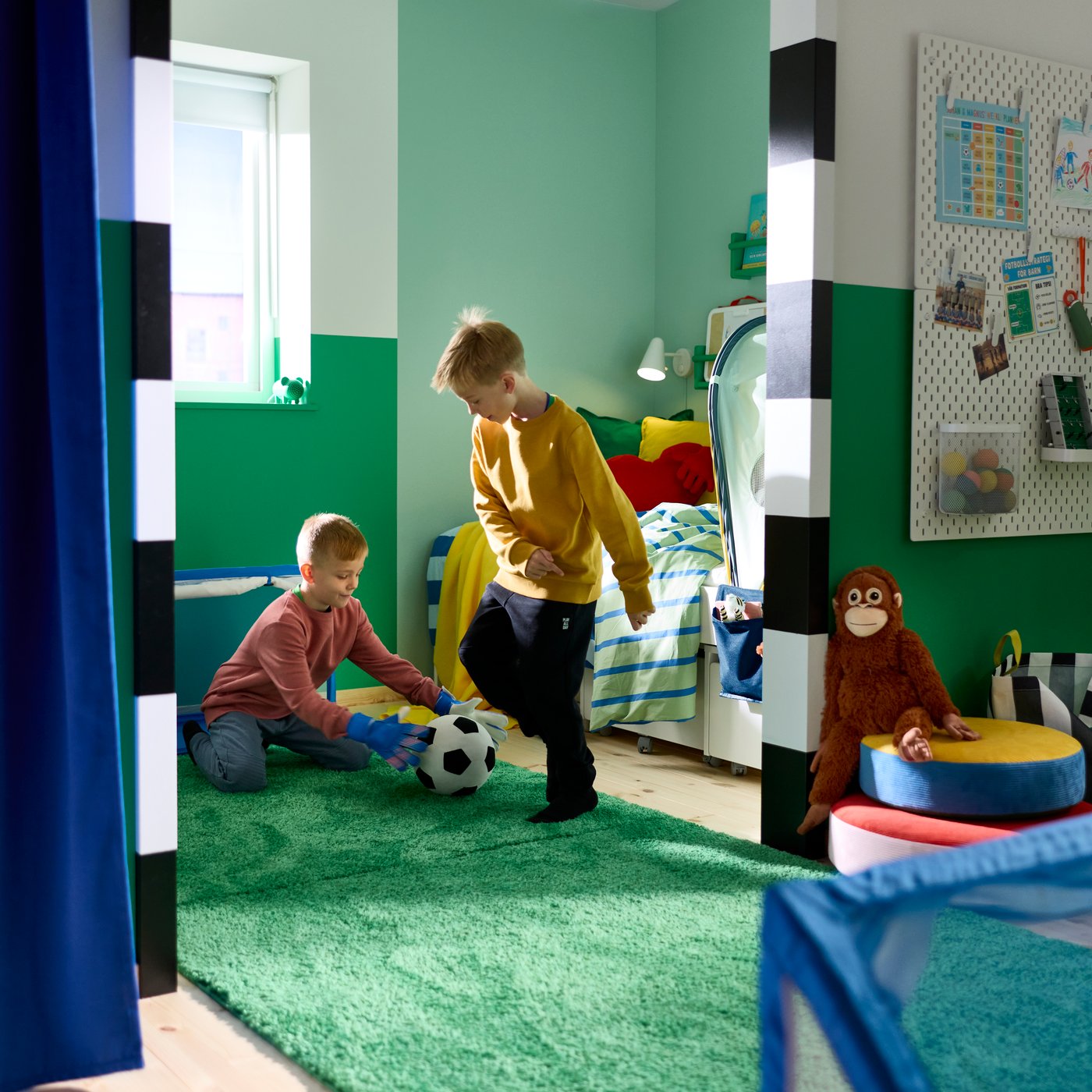 A green GREJSIMOJS portable Bluetooth speaker, shaped like a mouse, stands on a windowsill as two children play football.