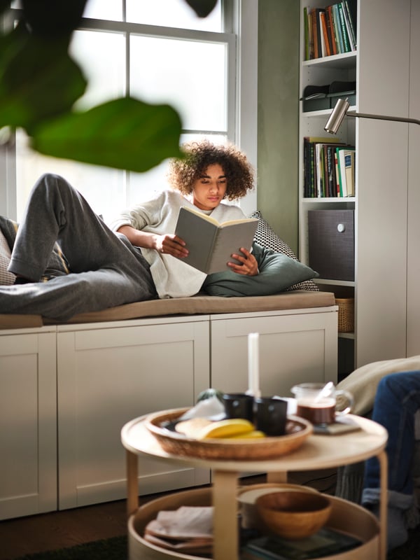 A teen reading while lying on a PLATSA storage bench in front of a white BILLY bookcase, filled with books and a speaker.