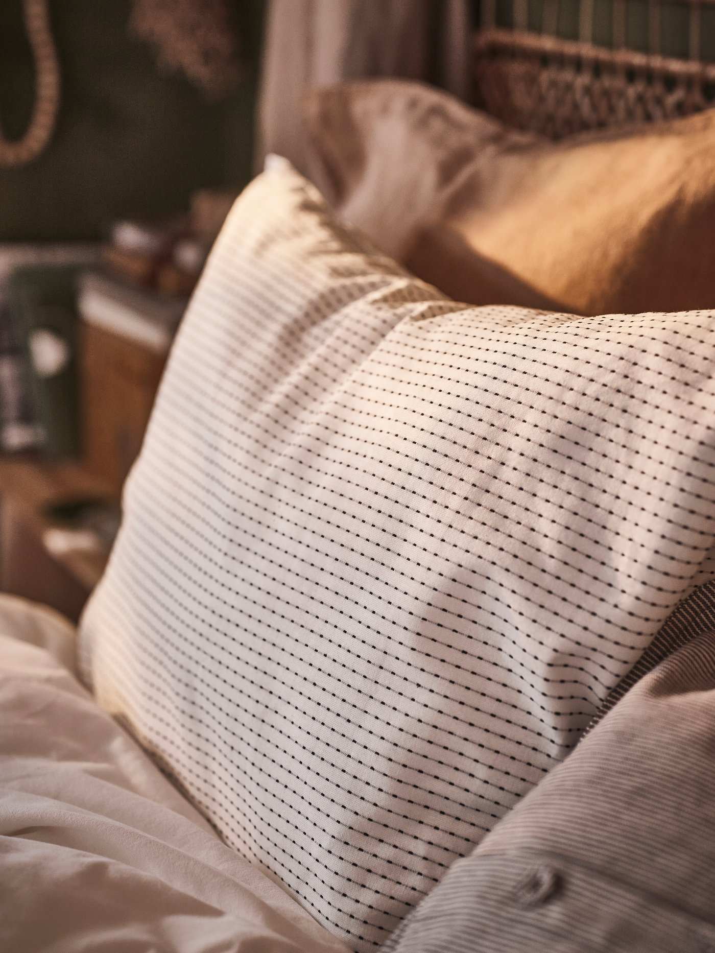 A white/dark grey TÅGVECKLARE pillowcase at the top of a bed in front of a pillow covered in a dark beige pillowcase.