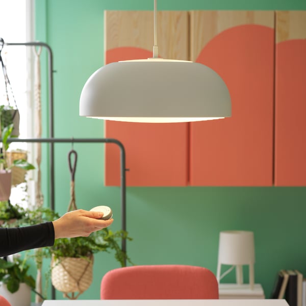 Ruka ispružena prema visećoj biljci u modernoj prostoriji sa zidovima pastelnih boja. Ruka drži mali, okrugli predmet, verovatno kamen ili ukrasni predmet. U pozadini je delimični pogled na nameštaj i rasvetna tela koja doprinose savremenom dekoru prostora.