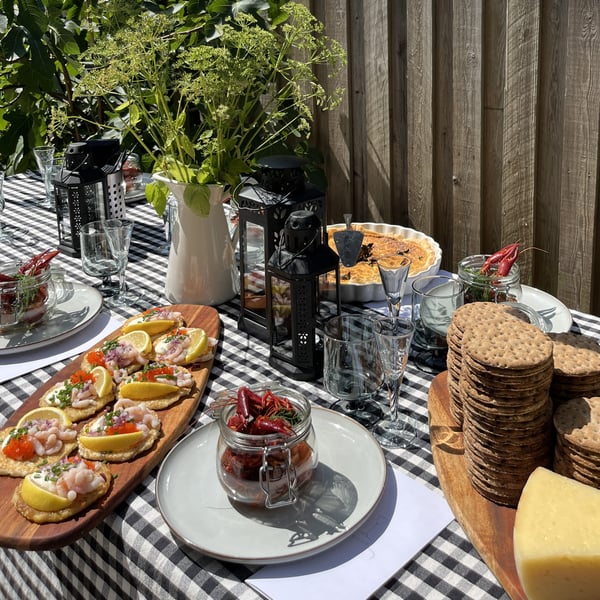 Utomhusbord är dukat inför kräftskiva med svartvit rutig duk, räkor på citron, ost, kex, inläggningar, glas och gröna dekorationer – perfekt för en somrig måltid i trädgården.