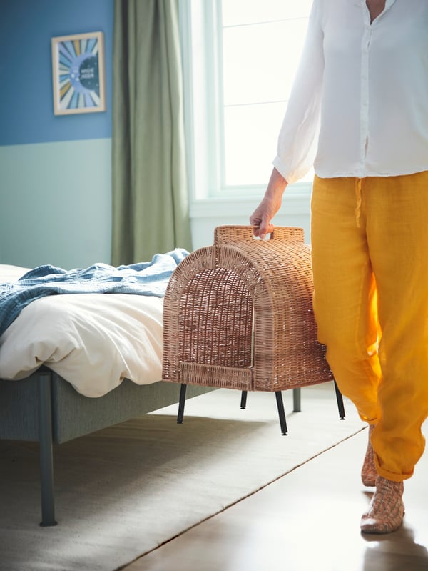 A person walking in the bedroom holding an IKEA UTSÅDD rattan cat house on legs. 