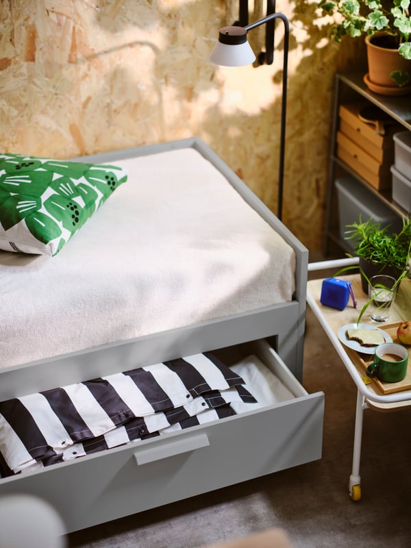 An open drawer of a grey BRIMNES day bed with 2 drawers showing folded black/white striped SLÅNHÖSTMAL bed linen inside.