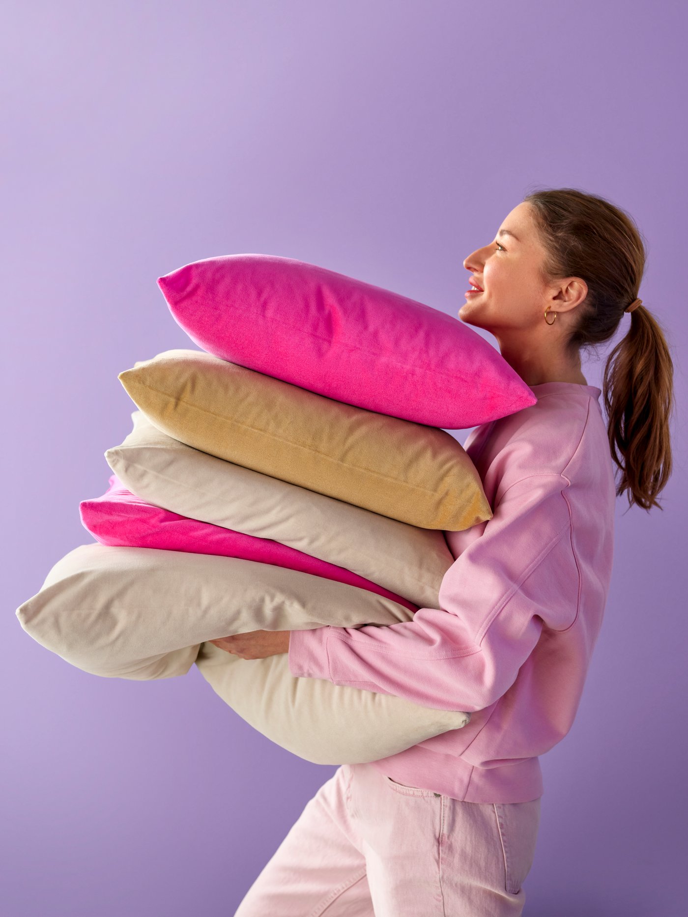 A person in profile holding a pile of cushions with SANELA cushion covers in bright pink, beige-yellow and light beige.