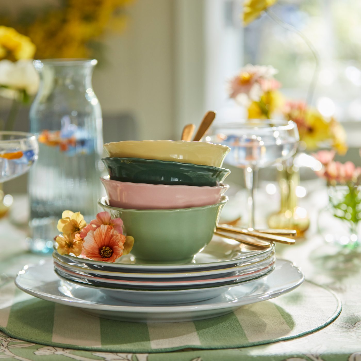 A stack of UPPLAGA bowls in yellow, green, and pink, placed on UPPLAGA side plates and a striped SMÖRFISK placemat.