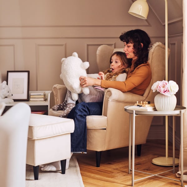 A child sits on an adult’s knee with a GREJSIMOJS soft toy on a STRANDMON wing chair. Beside them is a STRANDMON footstool.