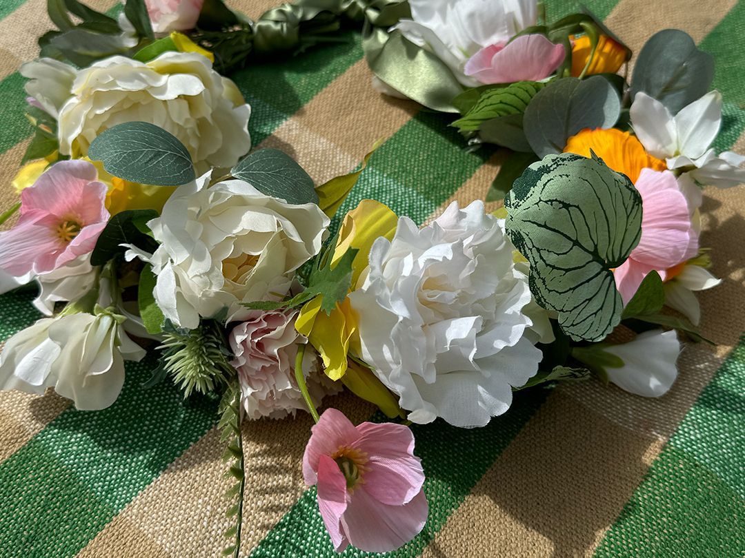 En egen midsommarkrans med konstgjorda blommor i olika färger ligger på en rutig duk.