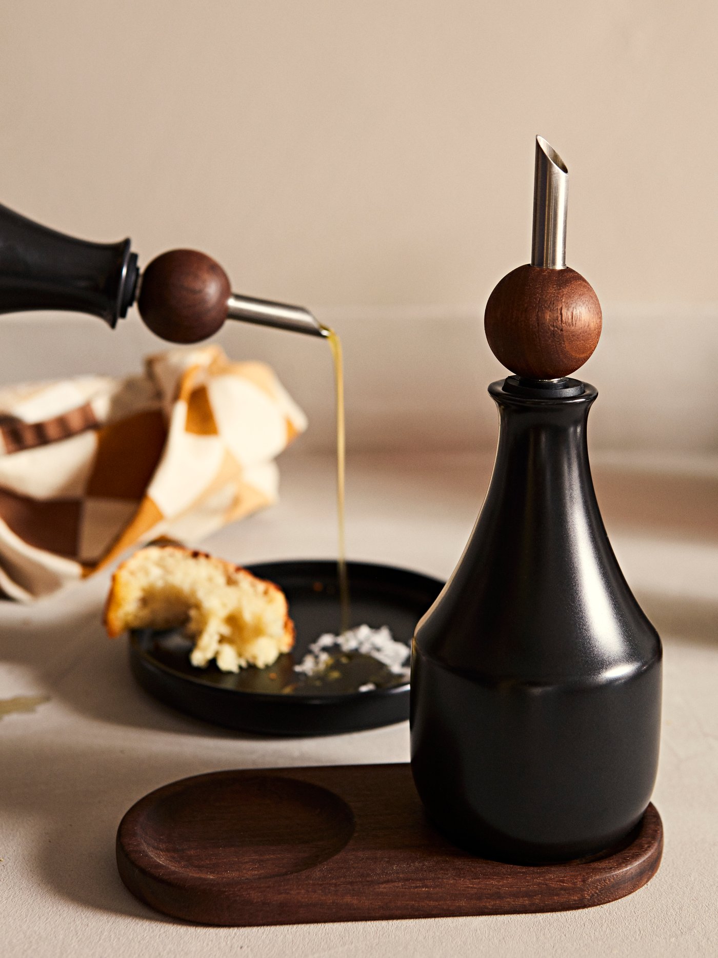 Olive oil being poured onto a KRÖSAMOS small plate with bread and salt. Another KRÖSAMOS bottle with pourer lid is in front.
