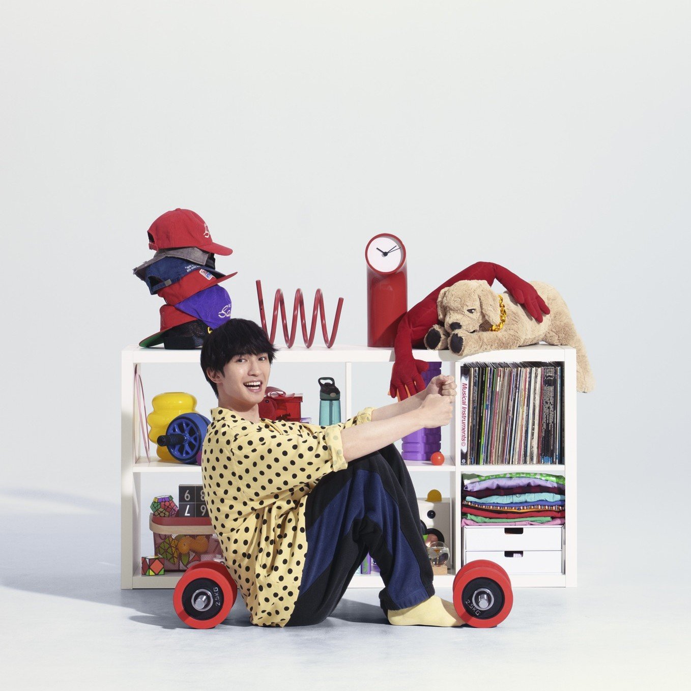 A person sitting before white storage shelves with a plush dog on their shoulder, hats, a clock, records and storage items.