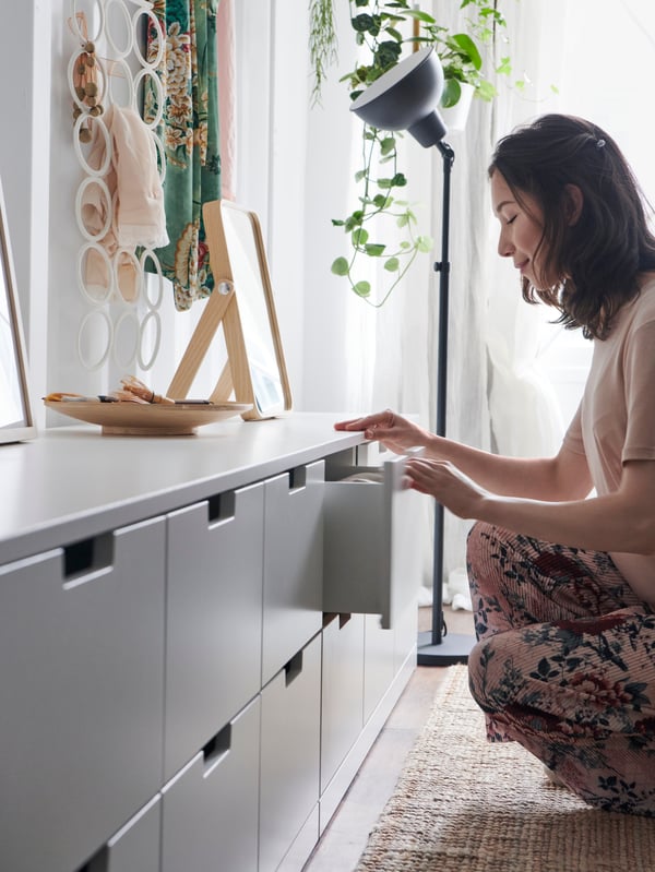 Dans une chambre scandinave, une jeune femme ouvre une commode blanche. Des accessoires déco est bois clair, des plantes vertes tombantes et un lampadaire noir créent une déco typiquement nordique.