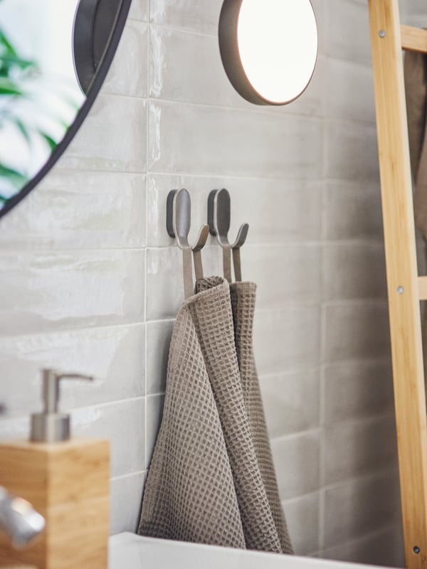 A softly lit bathroom with two light grey/brown towels hanging on two BROGRUND hooks on the wall near a soap dispenser.