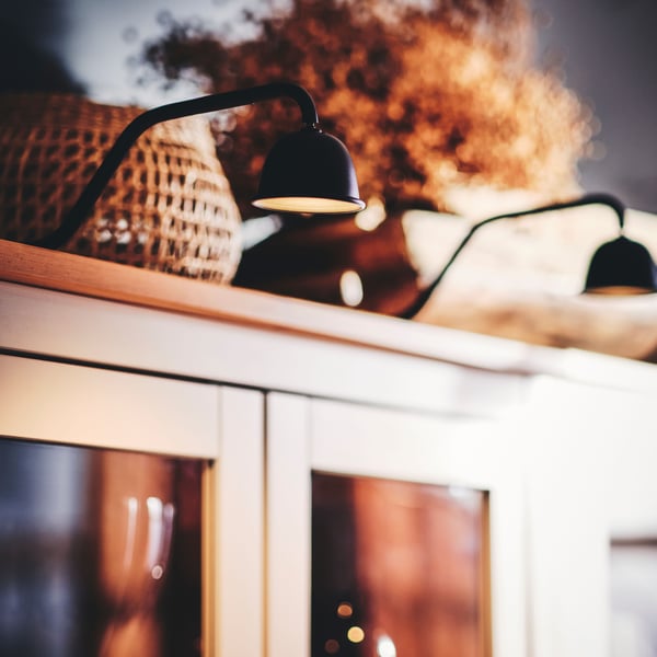 Two lamps of the TVÄRDRAG cabinet lighting in black illuminate a bookcase with glass doors and decorative times on top.