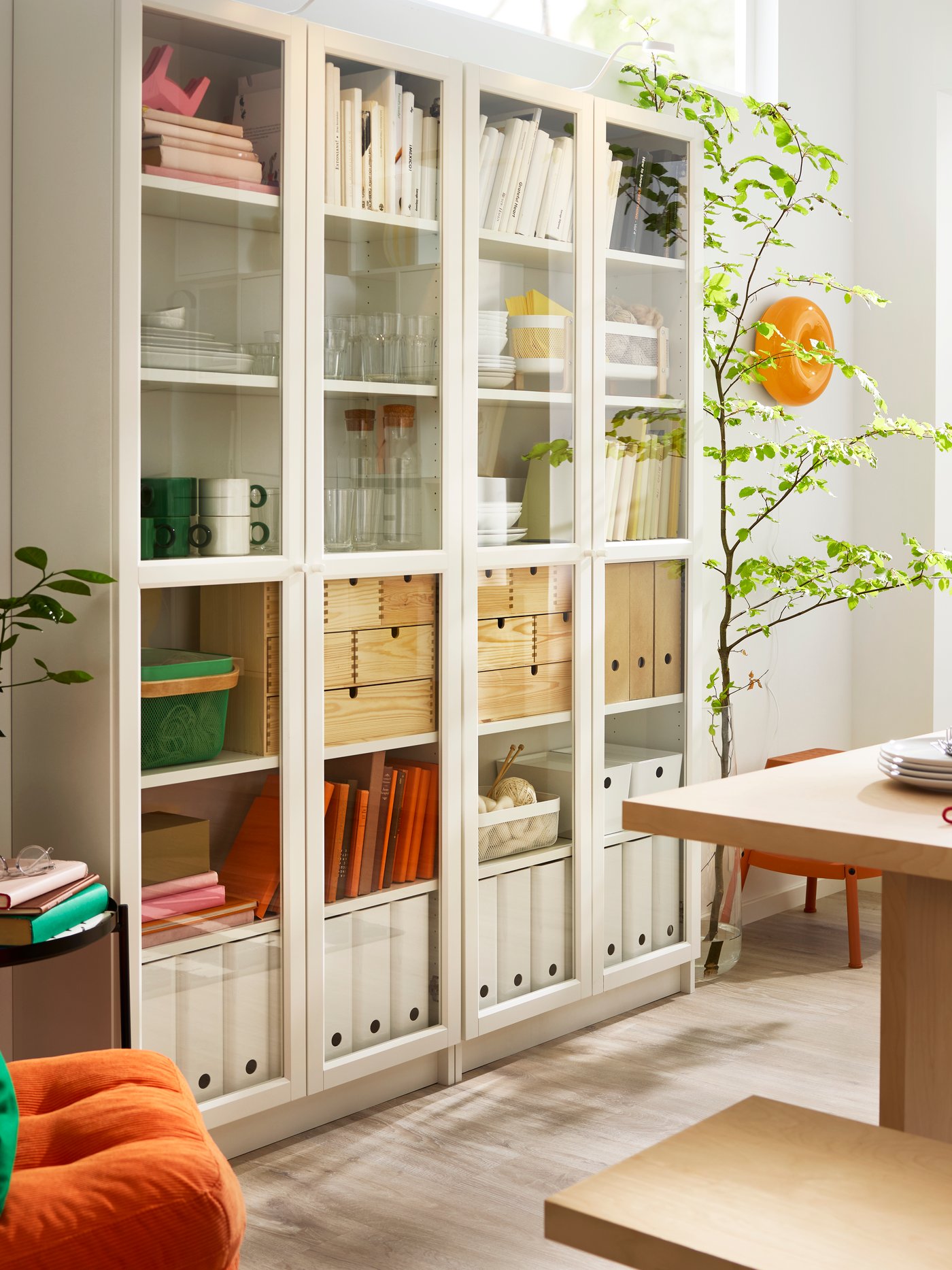 White BILLY bookcases with glass doors, placed next to a table, have binders, boxes, glasses and mugs placed on the shelves.