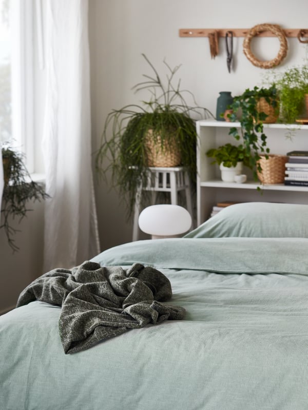 Una cama hecha con ropa de cama BERGPALM de rayas verdes. Libreros blancos BILLY a lo largo de la cabecera están llenos de plantas verdes.