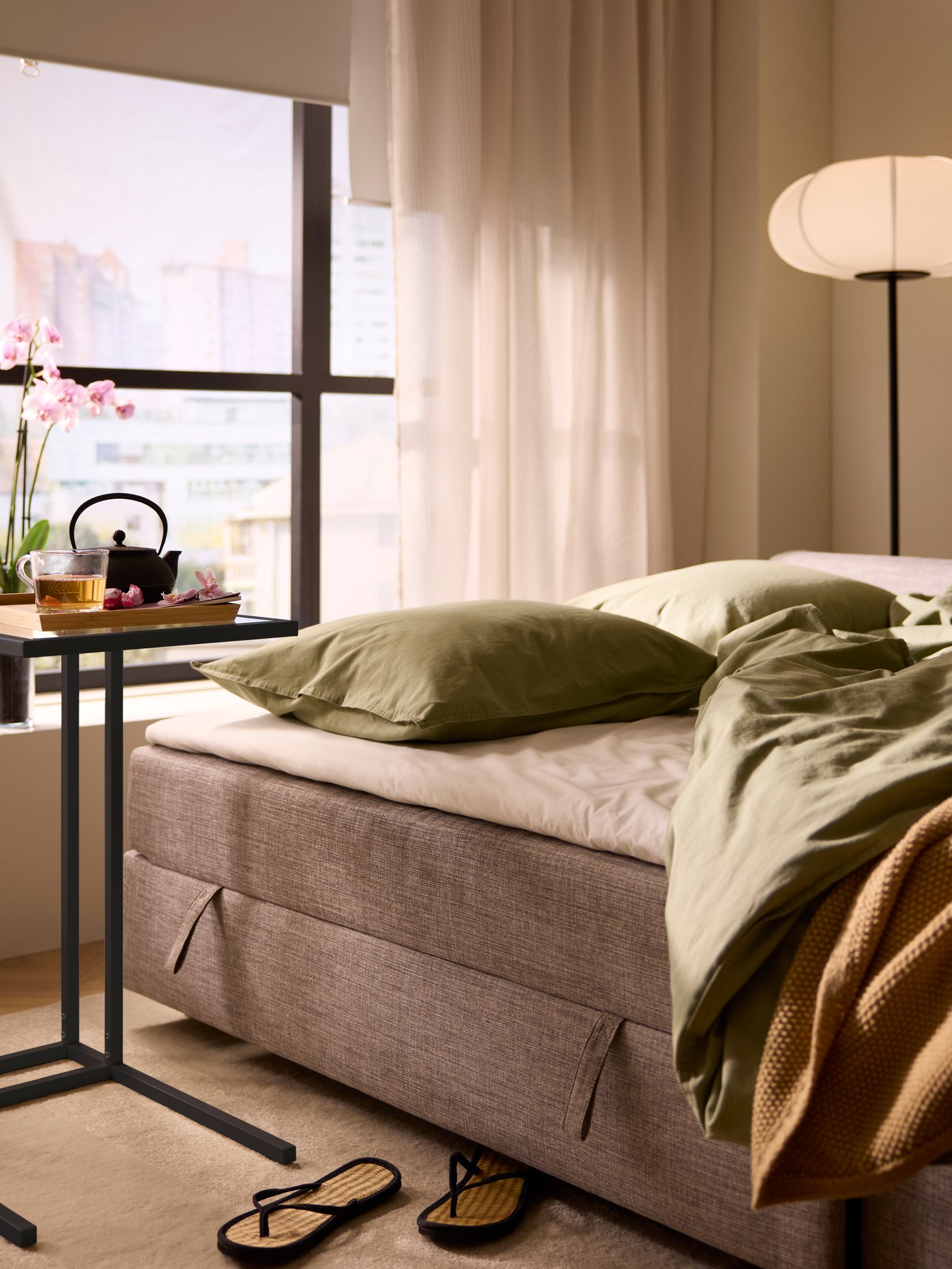 A tray with tea accessories on a VITTSJÖ laptop stand by a bed-mode FRIHETEN sofa-bed, in turn standing by a sunlit window.