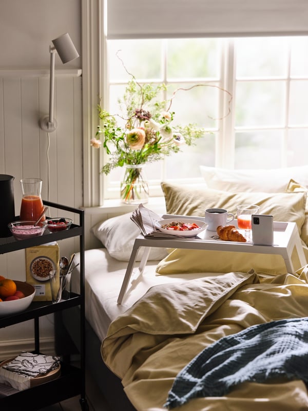 Carrello NISSAFORS nero con prodotti per la colazione accanto a un vassoio con colazione completa su un letto con biancheria ÄNGSLILJA.
