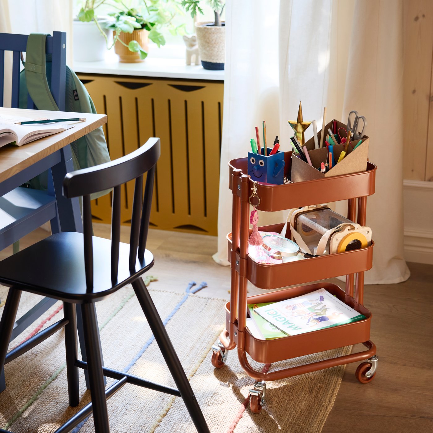 A red-brown RÅSKOG trolley holding pencils, books and other things, stands beside an AGAM junior chair on a RAKLEV rug.